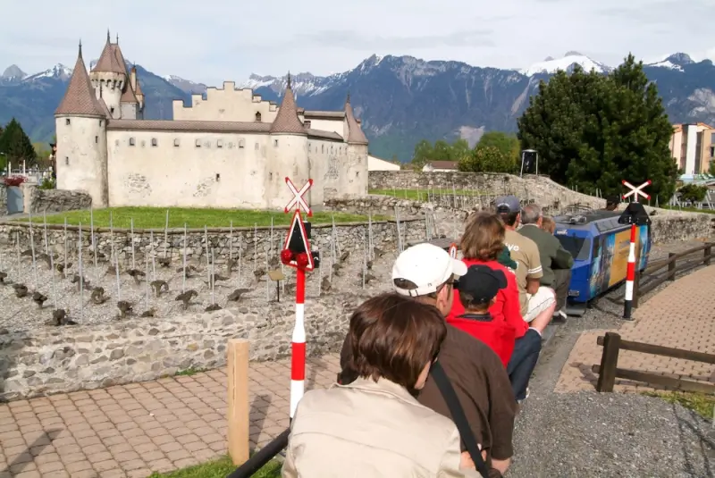 Families riding miniature train through detailed Swiss landscape with castle replica and Alpine mountain backdrop at Swiss Vapeur Parc