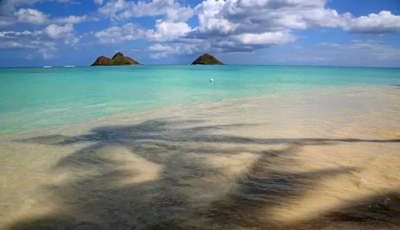 View of Mokulua Islands from Lanikai Beach with turquoise and aqua waters, shallow sandy shoreline, and dramatic cloudy sky in Oahu, Hawaii