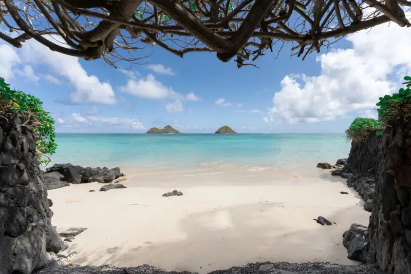 Secluded white sand beach framed by volcanic rock walls and tropical vegetation with Mokulua Islands visible across calm turquoise waters in Hawaii