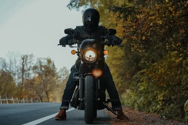 Motorcyclist in black riding gear sitting on stationary motorcycle on roadside with autumn trees in background