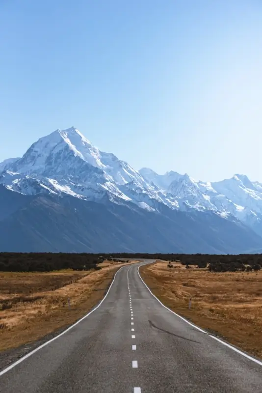 Scenic highway leading toward snow-capped Mount Cook with golden grasslands and clear blue sky in New Zealand