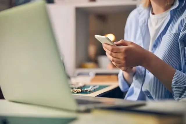 Person browsing jewelry on their phone while sitting at a desk with a laptop, suggesting a customer shopping for diamonds online.