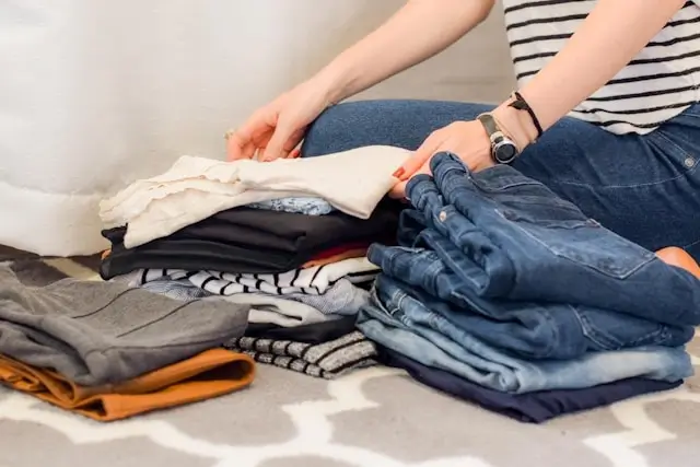 Woman in striped shirt organizing and folding clothes including jeans, shirts, and sweaters on bed while preparing for trip