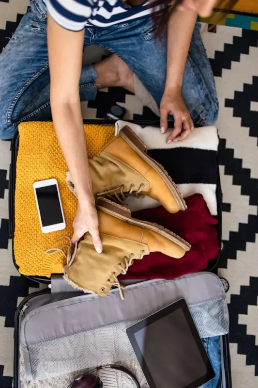 Traveler packing open suitcase with clothing, tan boots, yellow knit sweater, smartphone, and tablet on patterned rug