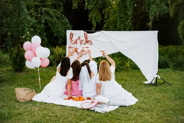 Bride and friends toasting champagne at outdoor bachelorette party picnic with pink balloons