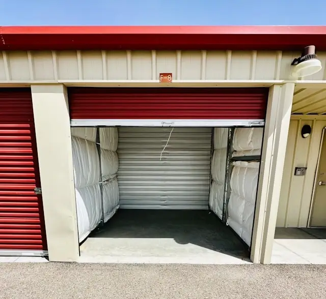 Open storage unit with burgundy roll-up door revealing interior packed with white wrapped mattresses and furniture along the walls, demonstrating storage capacity and organization