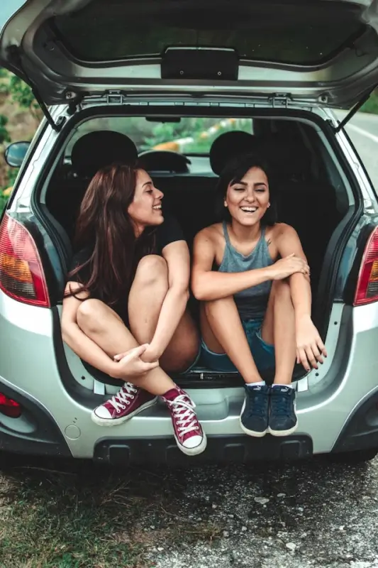 Two friends laughing together in car trunk during travel break, enjoying good company on long drive