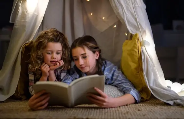 Two siblings reading a book together inside a cozy blanket fort with fairy lights