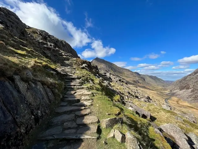 Stone hiking trail winding up mountainside with dramatic valley views under blue sky in Snowdonia National Park, Wales