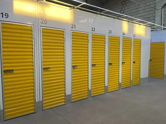 Row of bright yellow roll-up storage unit doors numbered 19 through 24 in a clean, well-lit indoor storage facility with white brick walls and concrete flooring