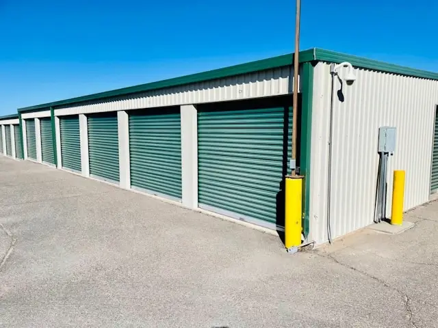 Exterior view of a self-storage facility with teal green roll-up doors along a white corrugated metal building under a blue sky, featuring yellow protective bollards near the entrance