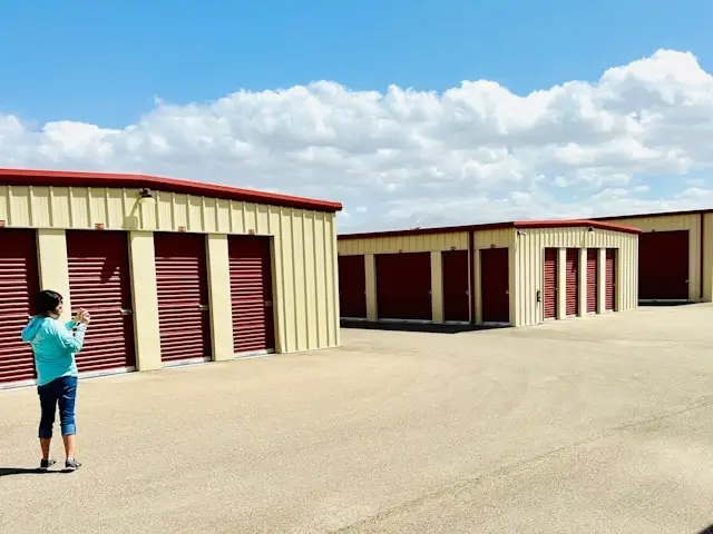 Person standing in front of outdoor storage units with burgundy roll-up doors on a beige metal building under a partly cloudy sky, viewing the available rental spaces