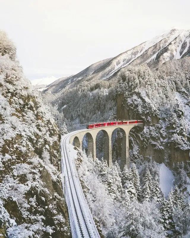 Red Swiss train crossing stone viaduct through snow-covered Alpine mountains in winter Switzerland