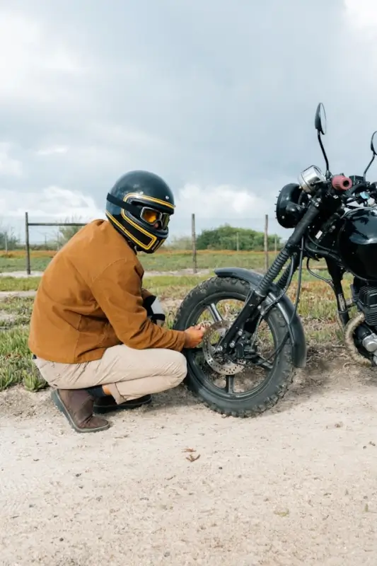 Motorcyclist in brown jacket and black helmet crouching beside motorcycle to inspect front tire and brake disc on dirt road