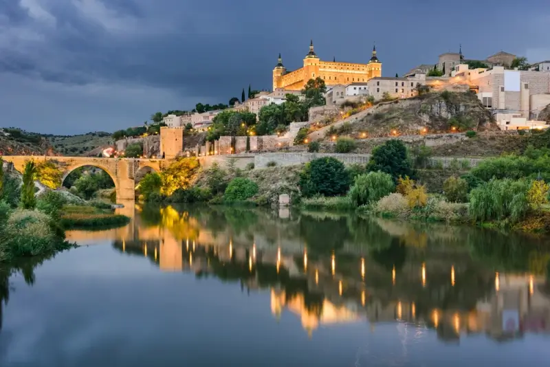 Toledo's Alcazar fortress and medieval cityscape illuminated at dusk, reflected in the Tagus River on a day trip from Madrid