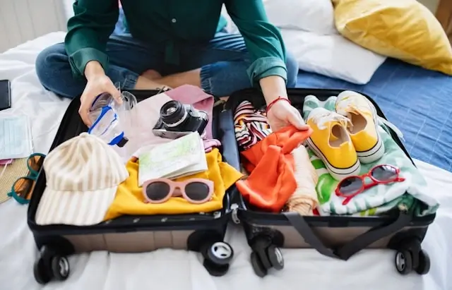 Person organizing open suitcase on bed with colorful clothes, camera, sunglasses, shoes, and travel gear neatly arranged