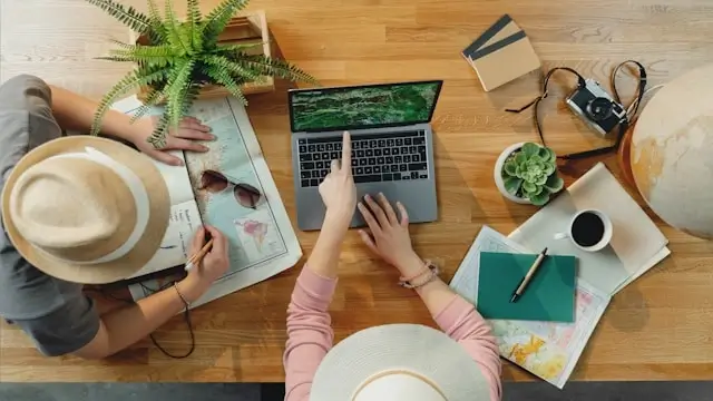 Two travelers planning their trip together at a wooden table with laptop, map, coffee, and travel accessories spread out