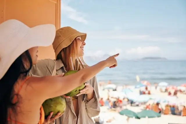 Two women travel with a companion enjoying a coastal view, pointing at the beach scene while wearing sun hats on a bright summer day