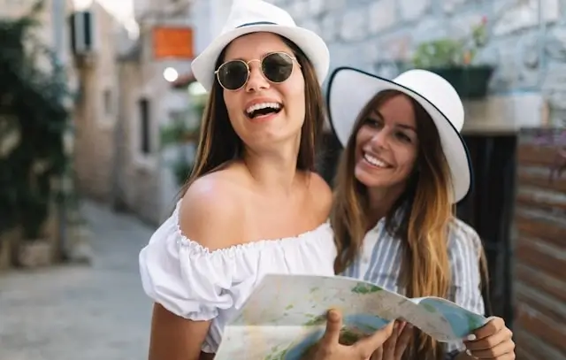 Two cheerful women laughing while holding a map and exploring a European city street wearing matching white sun hats