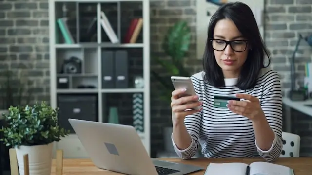 Young woman wearing glasses holding a phone and credit card while making an online purchase at her desk with a laptop open.