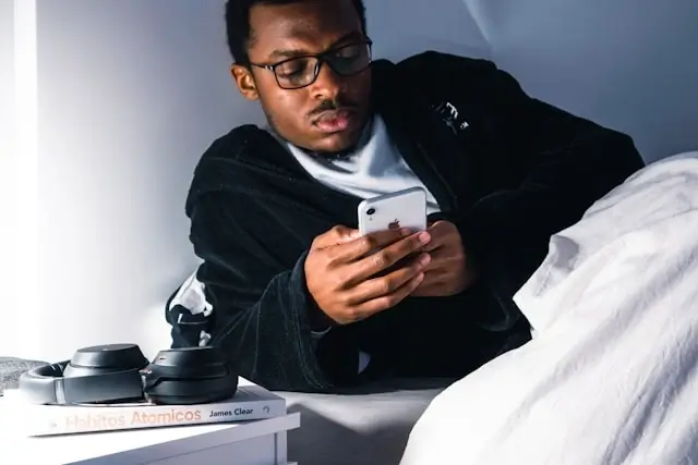 Man wearing glasses scrolling on phone in bed at night with books on bedside table, demonstrating habits that can interfere with quality sleep while traveling