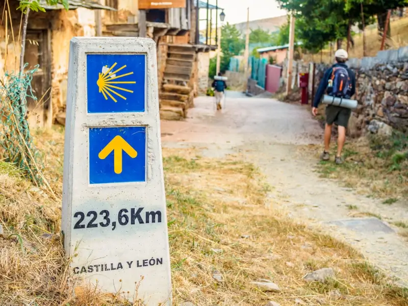 Pilgrim with backpack following yellow arrow waymarker showing 223.6km distance through Spanish village on Camino de Santiago tours