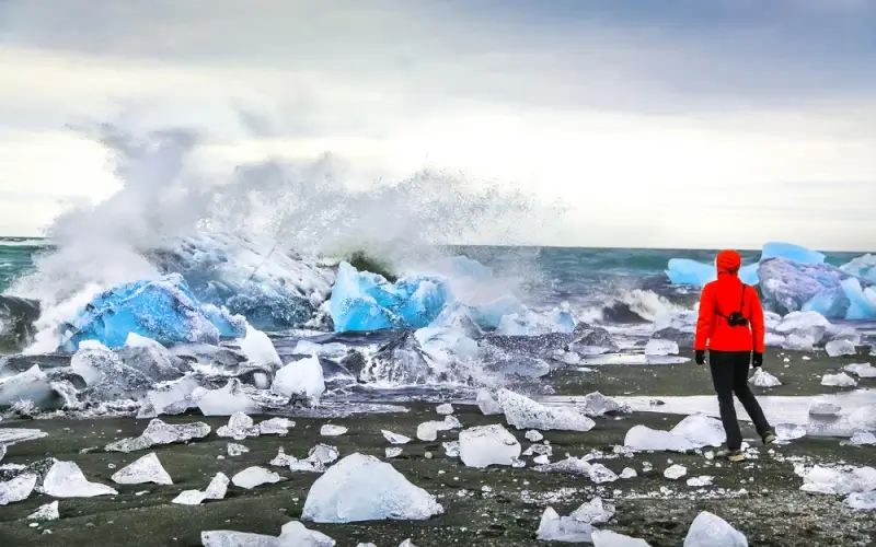 Woman in red jacket standing on black sand beach surrounded by ice chunks at Jokulsarlon glacial lagoon in Iceland with crashing waves