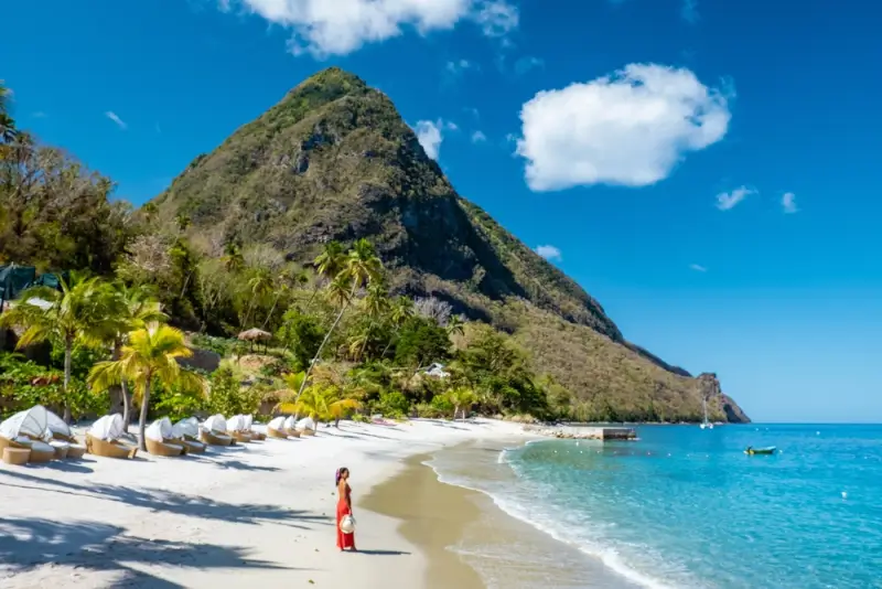 Woman in orange dress walking on white sand beach with turquoise water and Piton mountain in St. Lucia Caribbean