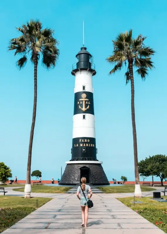Solo woman walking toward striped lighthouse framed by palm trees on sunny coastal day, embracing independent and leisurely journey