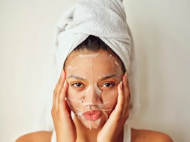 Woman with hair wrapped in towel applying face sheet mask as part of nighttime skincare routine before bed while traveling