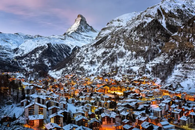 Aerial view of Zermatt village at dusk with illuminated chalets nestled in snowy Alpine valley below Matterhorn peak