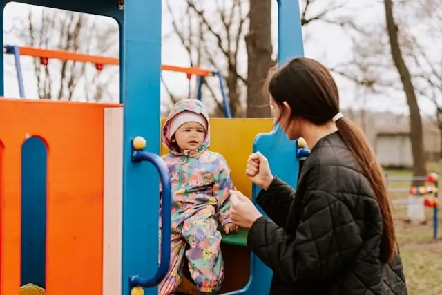 A young woman helping a toddler dressed in a colorful snowsuit while playing at a playground structure on a cool day.