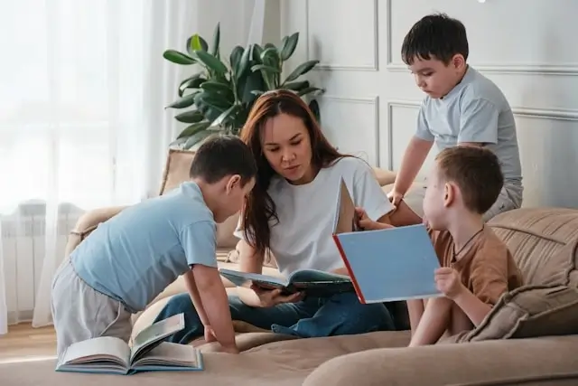 A woman sitting on a sofa reading books with three young children gathered closely around her in a cozy living room.