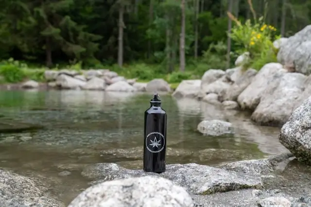 Black insulated water bottle with maple leaf logo on rocky riverbank surrounded by natural scenery