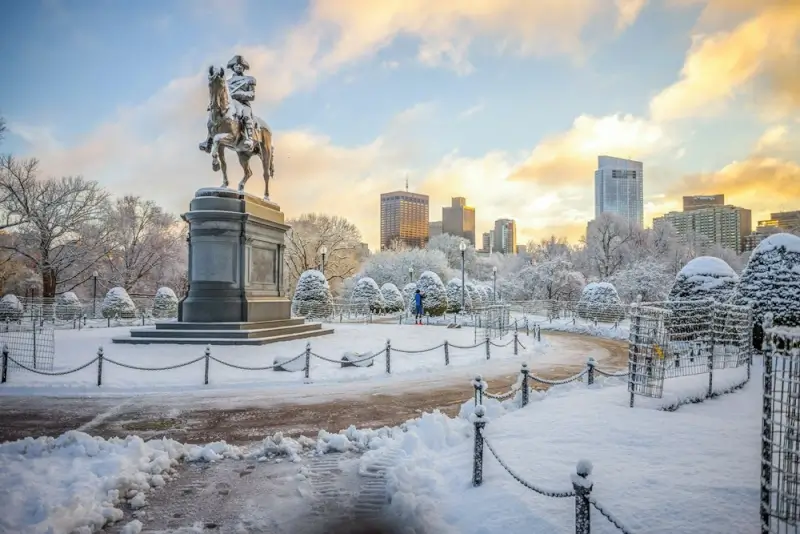 Snow-covered Boston Public Garden with George Washington statue and frost-covered trees against the city skyline