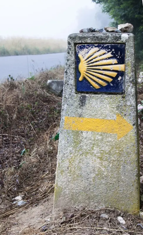 Ancient stone milestone with yellow scallop shell symbol marking pilgrimage trail to Santiago through misty Spanish countryside