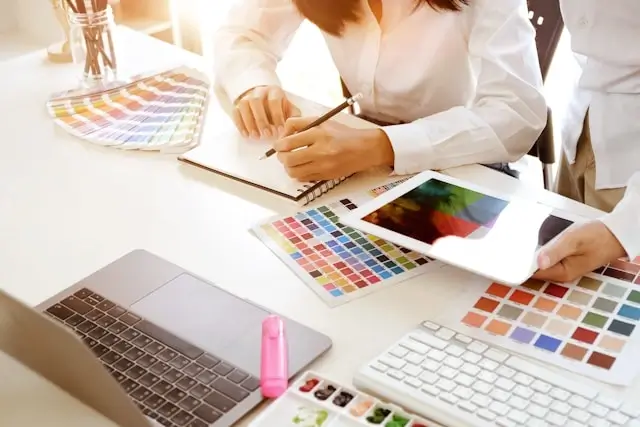 Designer selecting brand colors from multiple palette swatches with laptop and notebook on desk