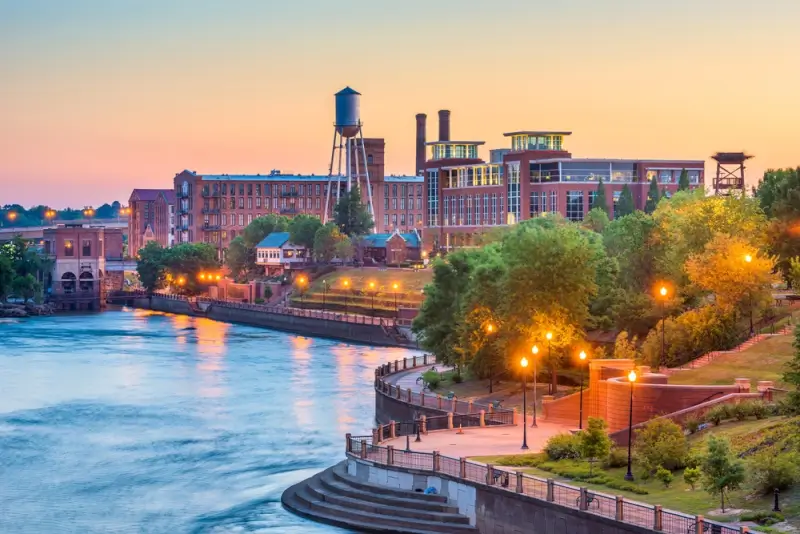 Columbus Georgia downtown skyline at sunset with historic brick mill buildings along the Chattahoochee River and illuminated Riverwalk pathway