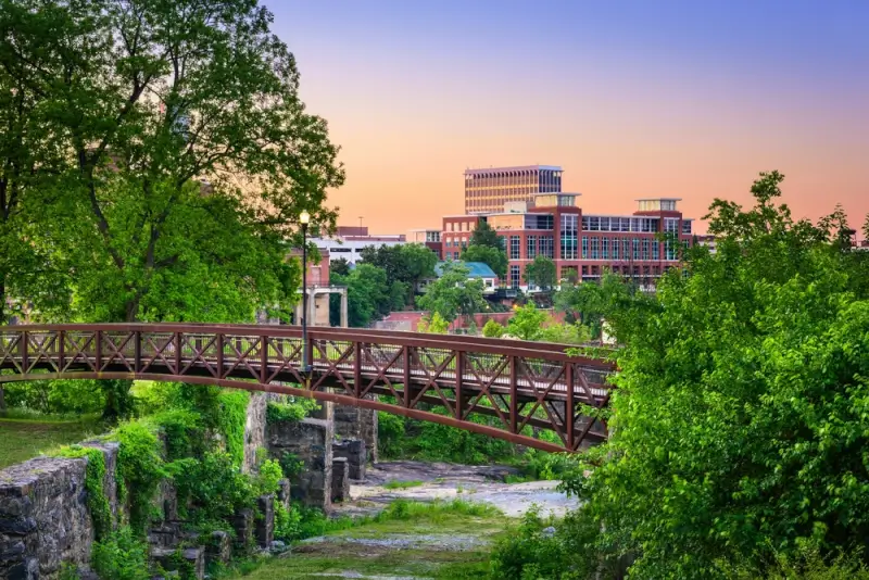 Wooden pedestrian bridge over moss-covered stone ruins in Columbus park with downtown buildings visible through lush green trees at twilight
