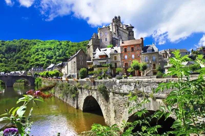 Traditional stone village in Pyrenees valley with medieval castle, old bridge crossing calm river, and lush green hillside under blue sky