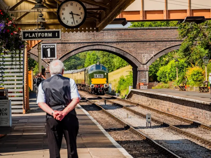 Heritage steam train arriving at Platform 1 station in the Cotswolds with railway worker watching from platform