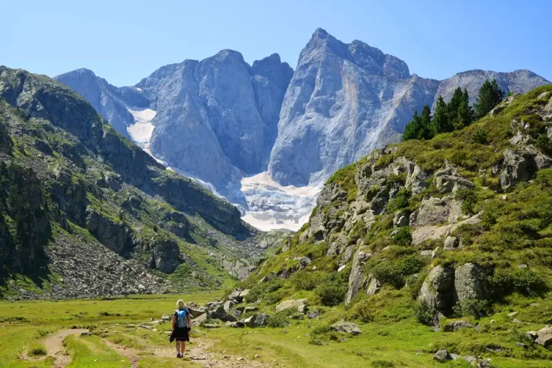 Hiking in the Pyrenees along green valley trail with hiker walking toward dramatic jagged peaks and glacier under clear blue sky