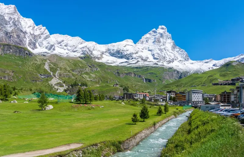 Aosta Valley village with flowing stream through green meadows and snow-capped peaks under blue summer sky in Italian Alps