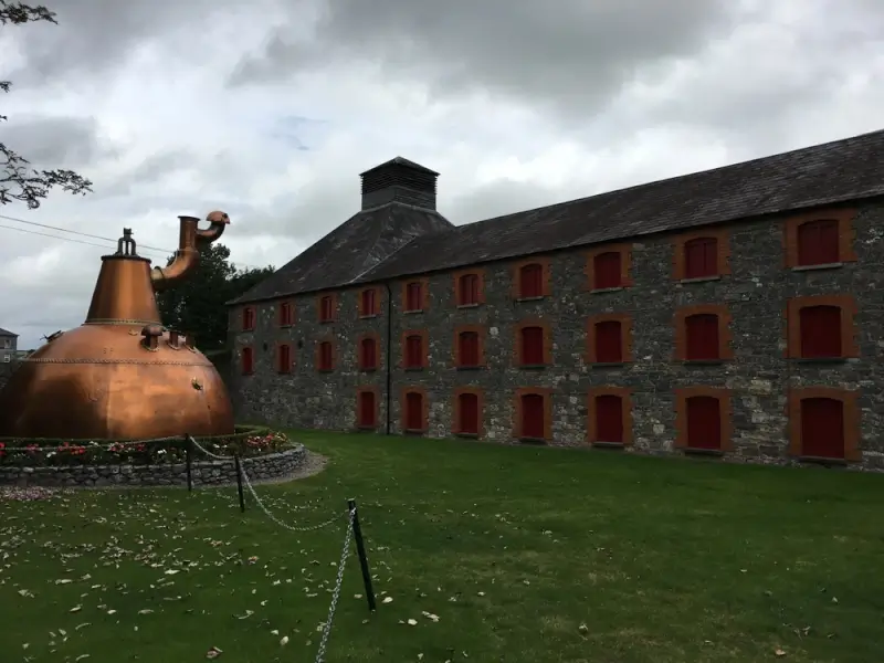 Historic stone distillery building with red-trimmed windows and traditional copper pot still on display, a popular stop on whiskey tours in Ireland