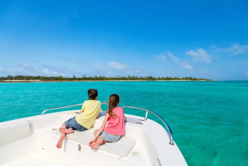 Two children sitting on bow of private boat in turquoise tropical waters with island coastline in distance