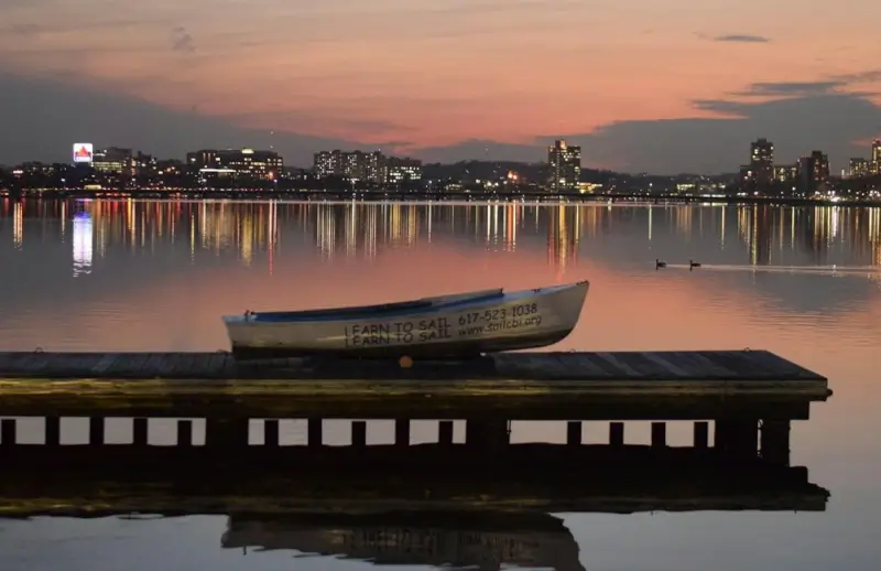 Learn to sail boat resting on dock at Charles River Esplanade with Boston skyline reflected in water at sunset