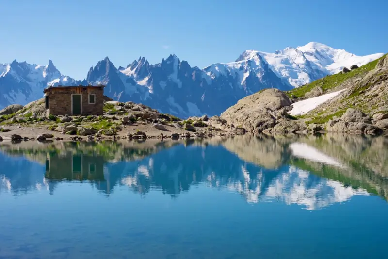 Mont Blanc massif and jagged peaks reflected in tranquil alpine lake with stone refuge on rocky shore