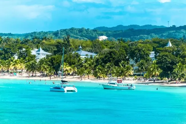 Turquoise waters and white sand beach at Negril, Jamaica with lush green mountains and boats in the bay