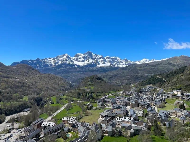 Mountain village nestled in green valley with snow-capped Pyrenees peaks rising dramatically in background under bright blue sky