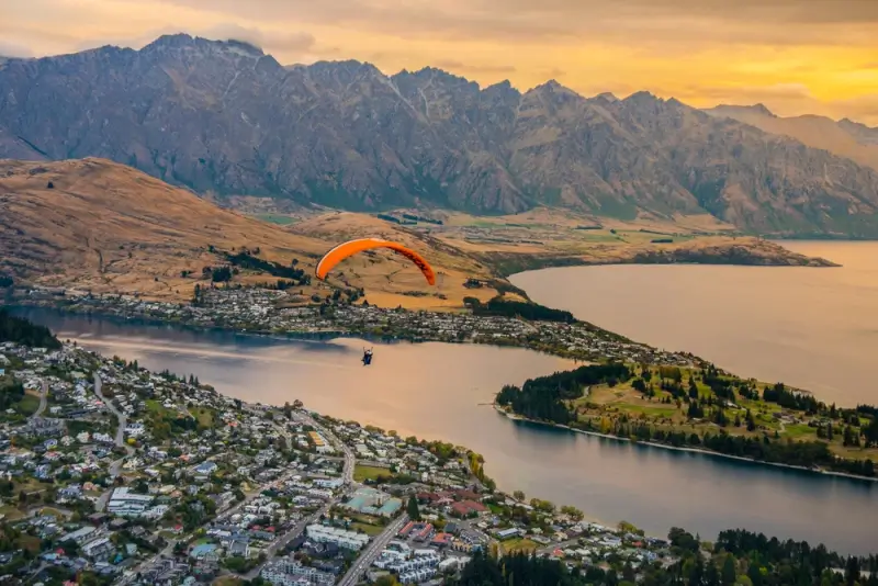 Sky adventures with paraglider gliding over Lake Wakatipu and Queenstown town with the Remarkables mountain range at sunset